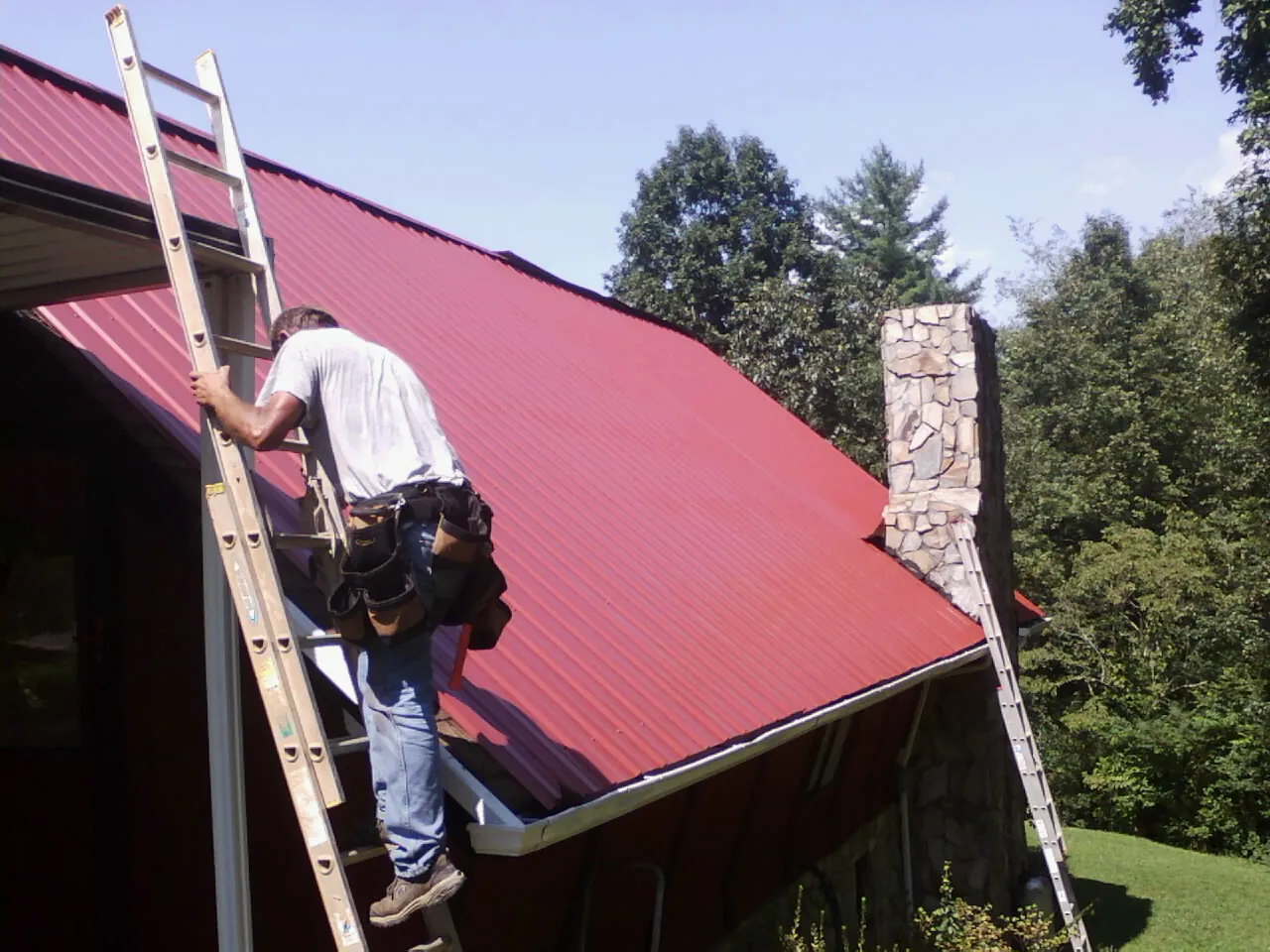 Roofer on a ladder beside a red metal roof and stone chimney — Soffit & Fascia Repair by Roofing Brothers in Rock Port